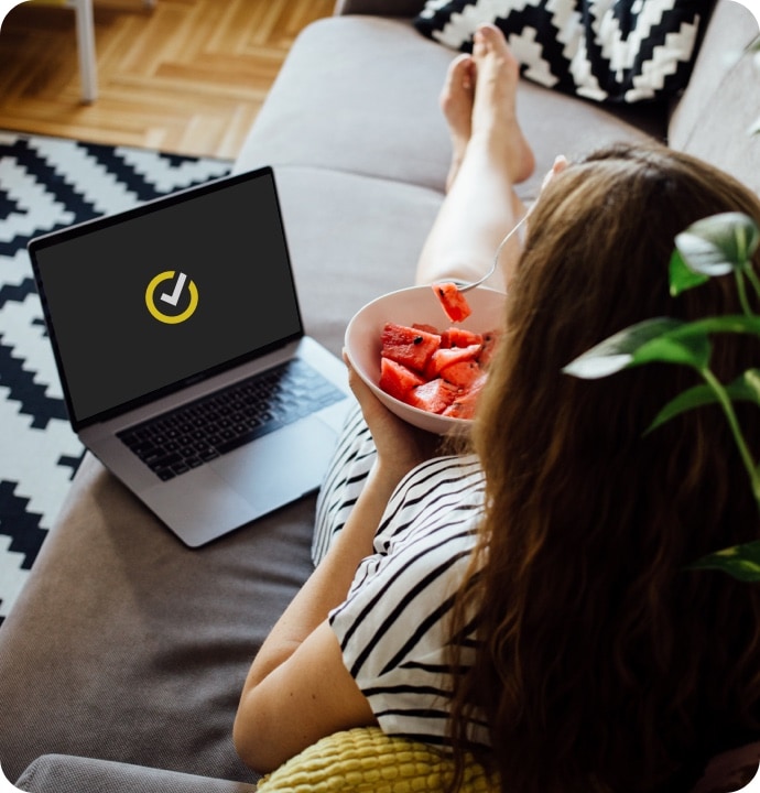 Kanepede uzanarak bir kase meyve yerken yanında Norton logosunu gösteren dizüstü bilgisayarlı kadın